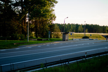 A quiet countryside bus stop sits alone beside a multi-lane highway, surrounded by trees and lit by golden sunset light.