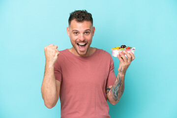 Young caucasian man holding a bowl of fruit isolated on blue background celebrating a victory in...