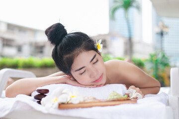 A woman lying on a massage bed with her eyes closed, with flowers and spa stones nearby. Represents...