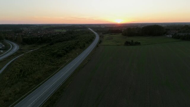Sunset sunrise gold hour over farmland fields and forest area split by traffic road with cars moving along. Residential buildings in the distance and strong orange and blue hues.