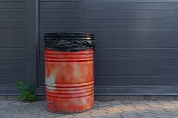 A red garbage barrel with a black bag inside stands near the wall of a modern building. Background. Street. Blank.