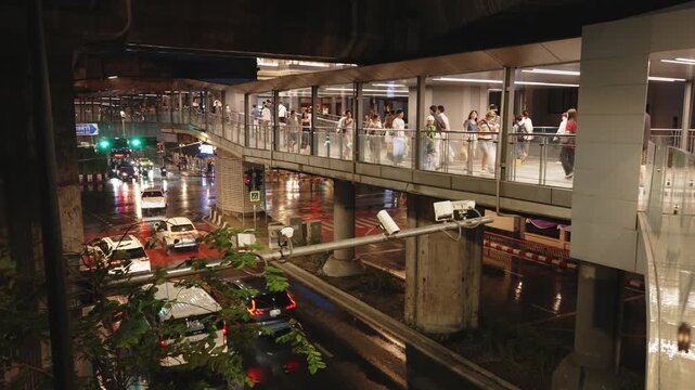 Pedestrian skywalk crowded with people above rainy night traffic near Siam station