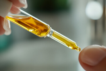 Close up of gloved hand holding glass dropper, releasing a golden viscous liquid drop. Natural plant extract or cosmetic oil being precisely applied.