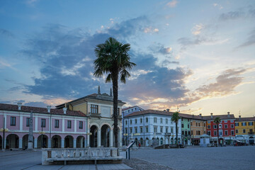 Malowniczy plac miejski z charakterystyczną palmą na pierwszym planie, otoczony kolorowymi budynkami o pastelowych elewacjach. Na niebie widoczne są chmury i delikatne światło zachodu słońca, tworzące © Alina