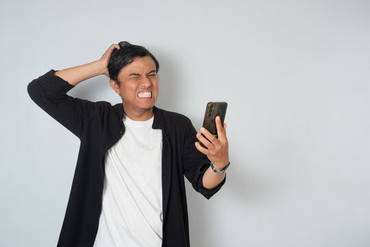 Asian man looking at smartphone with confused expression, scratching his head in frustration. Concept of bad news, stress, disappointment, and problem with mobile device on white background.