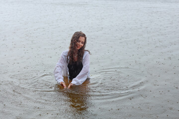 Fototapeta premium Beautiful young brunette woman in black swimsuit and white shirt standing in water of the sea near sandy shore in rainy weather