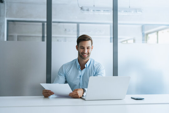 Smiling latin business man salesman working on laptop computer reading financial contract document in office workplace. Young manager businessman specialist doing paperwork analysing on pc. Copy space
