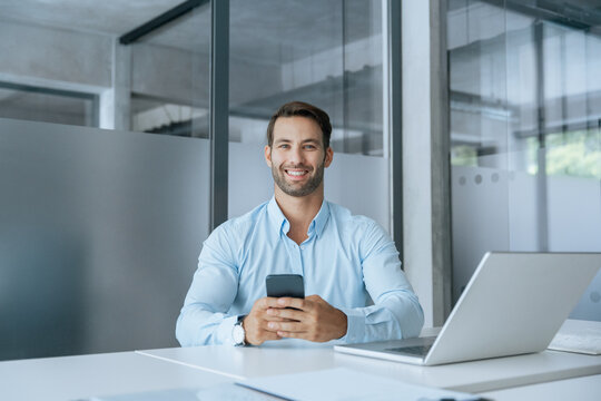Middle-age manager man ceo using cell phone mobile app, laptop. Smiling Latin young businessman holding smartphone in business office working crypto online, looking at camera portrait. Copy space