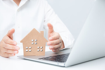 Businessman using both hands to protect a house model with a laptop beside it. Concept of property protection, real estate security, insurance, investment, and safe business planning