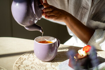 Woman pouring tea from a purple teapot into a matching cup, surrounded by pastries on a lace tablecloth, evoking elegance and calm morning rituals