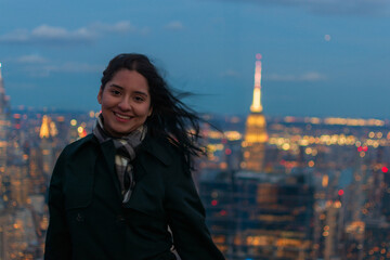 Smiling young Hispanic woman on New York City skyscraper rooftop with glowing Empire State Building at night