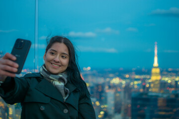 Smiling young Hispanic woman taking selfie with smartphone on New York City skyscraper rooftop at night with glowing Empire State Building in background