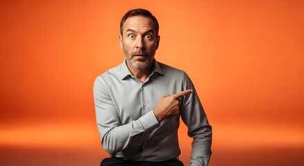 Fototapeta premium Astonished Man in Gray Shirt Points Right with Wide Eyes Against Vibrant Orange Gradient Background.