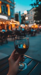 A wine glass filled with red liquid held by a person's hand is centered, with a blurred, cozy city backdrop at dusk