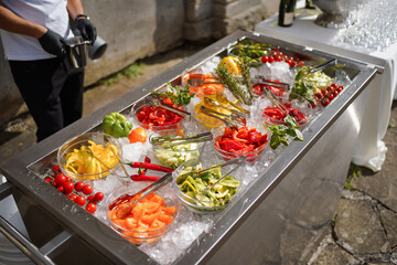 Fresh sliced vegetables on ice in glass bowls with tongs. Colorful peppers, tomatoes, herbs displayed for buffet or outdoor catering.