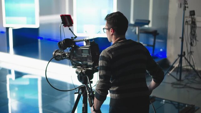 Camera operator adjusting professional video camera setup in modern studio. Focused on filming with studio lights and high-tech equipment, cables, and broadcast production tools visible
