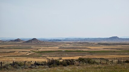 Expansive semi-desert landscape of Bardenas Reales in Navarre, Spain, features uniquely shaped hills and mesas. The terrain is predominantly tan and brown with patches of sparse vegetation. 
