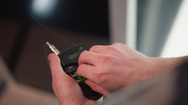 hands of technician open battery compartment of wireless audio transmitter inserting colorful rechargeable batteries inside handheld unit under bright studio lights during broadcast setup preparation