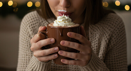 Woman sips hot chocolate with whipped cream in a brown mug wearing a cozy sweater indoors with lights