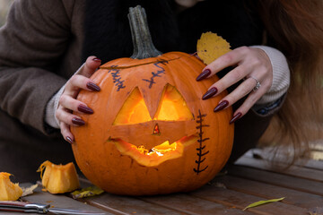 Close-up view of woman with red long sharp fingernails touching orange carved Halloween pumpkin with glowing face lying on wooden table in autumn park. Handheld video. Holiday's symbol theme.