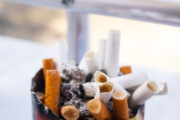 A detailed, close-up shot shows a cluster of burnt-out cigarettes and butts with their filters exposed, filling a rusty or old ashtray
