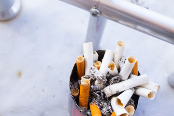 A detailed, close-up shot shows a cluster of burnt-out cigarettes and butts with their filters exposed, filling a rusty or old ashtray