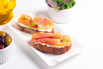 Two bruschettas with prosciutto, ricotta cheese and rucola on ciabatta bread. Olive and spices  decorated.