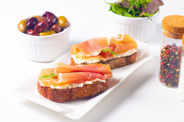 Two bruschettas with prosciutto, ricotta cheese and rucola on ciabatta bread. Olive and spices  decorated.