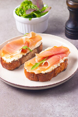 Two bruschettas with prosciutto, ricotta cheese and rucola on ciabatta bread. Olive and spices  decorated.