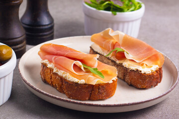Two bruschettas with prosciutto, ricotta cheese and rucola on ciabatta bread. Olive and spices  decorated.