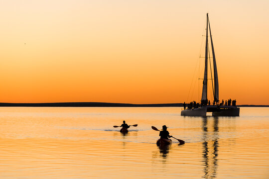 Kayakers paddle past a yacht at sunset on calm golden waters.