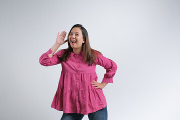 A cheerful young woman wearing a pink blouse smiling and holding her hand near her ear as if...