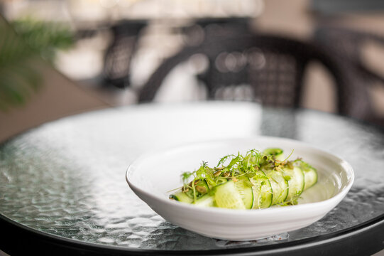 Fresh cucumber slices garnished with herbs in a white ceramic bowl on a glass table, outdoor setting, minimalistic food presentation, healthy vegetarian snack. - Powered by Adobe
