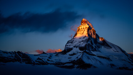 The matterhorn peak illuminated by the first light of dawn, with snowcovered slopes and dramatic clouds in the dark blue sky
