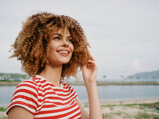 Portrait of a young woman with curly hair wearing a red striped shirt, standing outdoors by a calm...