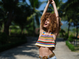 joyful young woman with curly hair wearing colorful striped crop top stretching arms outdoors,...