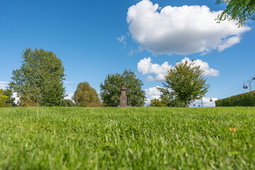 A green meadow with trees and a stone statue under a bright sky. Cable cars float in the distance among white clouds.
