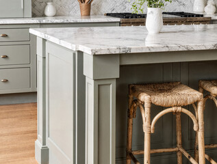 A sage green farmhouse-style kitchen island is finished in sleek white marble. Neatly placed rattan bar stools stand out, highlighting the textures of the marble and wood flooring. This creates a coz