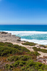 A breathtaking coastal view showcasing turquoise ocean waves crashing against rocky cliffs, surrounded by lush green shrubs and sandy terrain under a clear blue sky