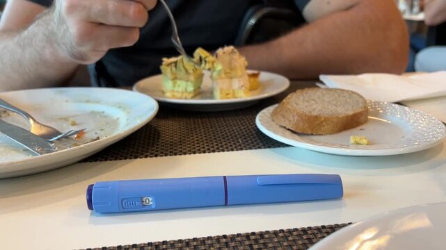 A man eats cake surrounded by empty food plates, with an Ozempic injection pen placed prominently in the front of the table. 