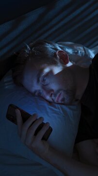 A sleepless young man lies awake in his bedroom at night, using his smartphone scrolling social media. Digital dependence and insomnia concept. vertical video