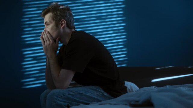A stressed man sits on the edge of a bed in a dark room at night, holding his head in distress. Depression, burnout or mental health problems. Copy space