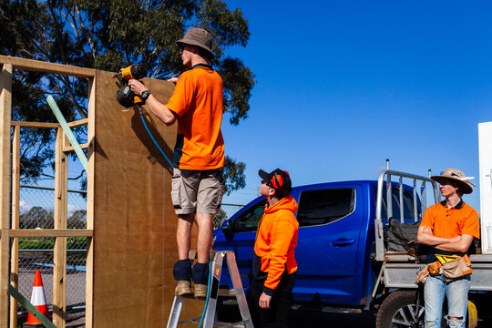 Three trade apprentice workers building frame of ticket booth using nail gun