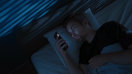Tired young man lies in bed at night, staring at his smartphone screen, doom scrolling through social media, unable to sleep struggling with sleep disorder or distraction by overthinking. Copy space - Powered by Adobe