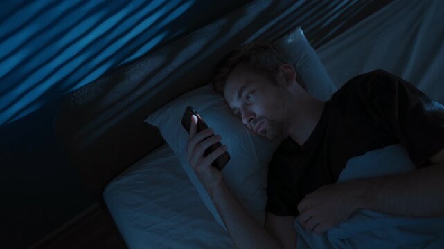 Tired young man lies in bed at night, staring at his smartphone screen, doom scrolling through social media, unable to sleep struggling with sleep disorder or distraction by overthinking. Copy space