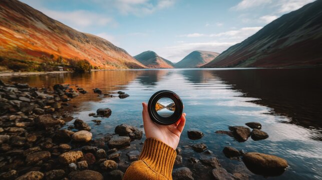 Through the camera lens held by a hand, a person frames a tranquil lake landscape with mountains reflected in it, seeking a future vision, inspired by the natural world, and continually searching for