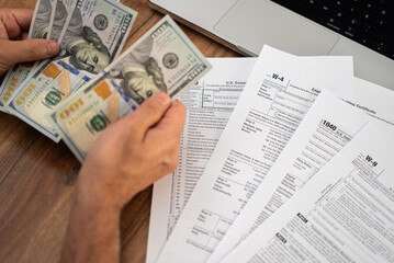 Male entrepreneur counts dollar banknotes preparing clear financial data for filling different tax forms mandatory for law-abiding taxpayers at workplace with laptop