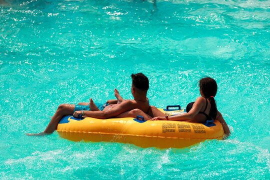 A couple enjoys a summer day together at a water park. They recline on a bright yellow inflatable tube, drifting lazily across the turquoise water - Powered by Adobe