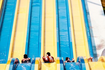 Three young boys are enjoying a sunny day at an Aqua Park. They are racing down the blue and yellow water slides, splashing in the water as they descend