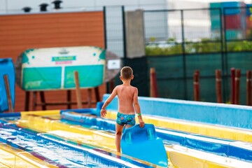 A shirtless young boy in swim trunks ascends a colorful water slide at a Water Park, carrying a...